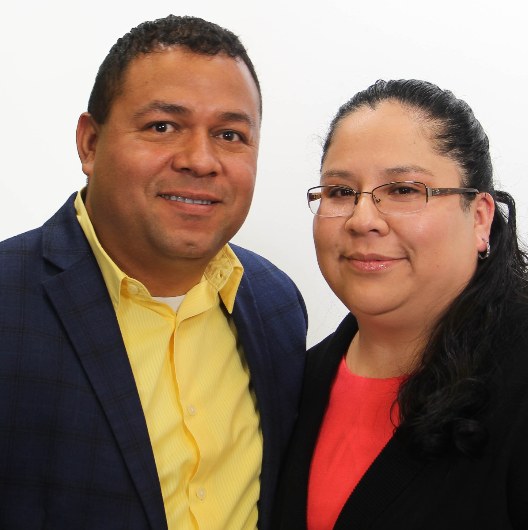 Two professionals posing together against a white background, man in navy blazer and yellow shirt, woman in red top and glasses